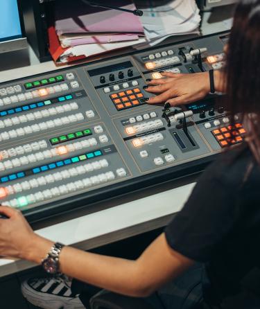 looking over the shoulder of a woman with black hair with her hands on a standard tv broadcast control board