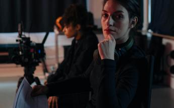woman film producer with dramatic lighting stares into camera while sitting in a director's chair with paperwork in hand. a camera and camera operator can be seen in the background