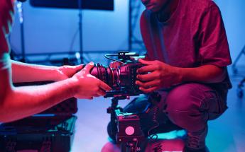 young black man assists a lens change on a cinema camera under pink and blue lighting