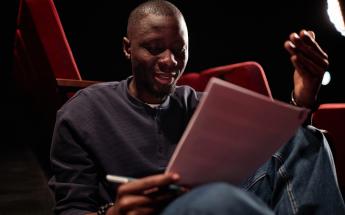 young black man sitting in dramatically lit environment reading and revising a film script while holding a pen
