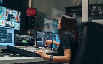 young woman with black hair sits in a chair while looking at a live video feed display along with a tv production control board