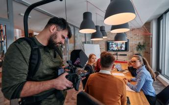 young man with pale skin and beard wearing green shirt operates a camera on an easyrig in a meeting room with employees chatting