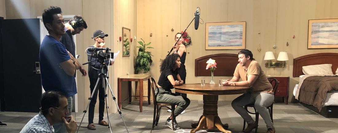 A student holds a boom mic on a pole over actors at a table. 