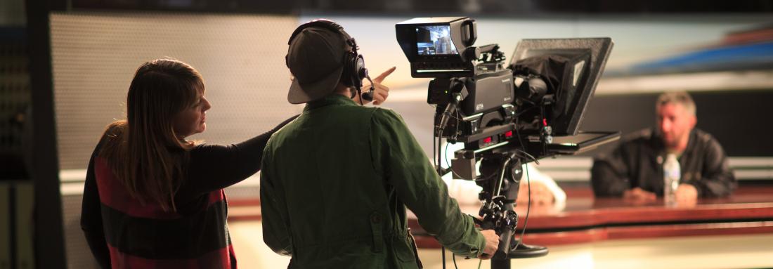 A director points to the camera monitor. A newsperson in the background.